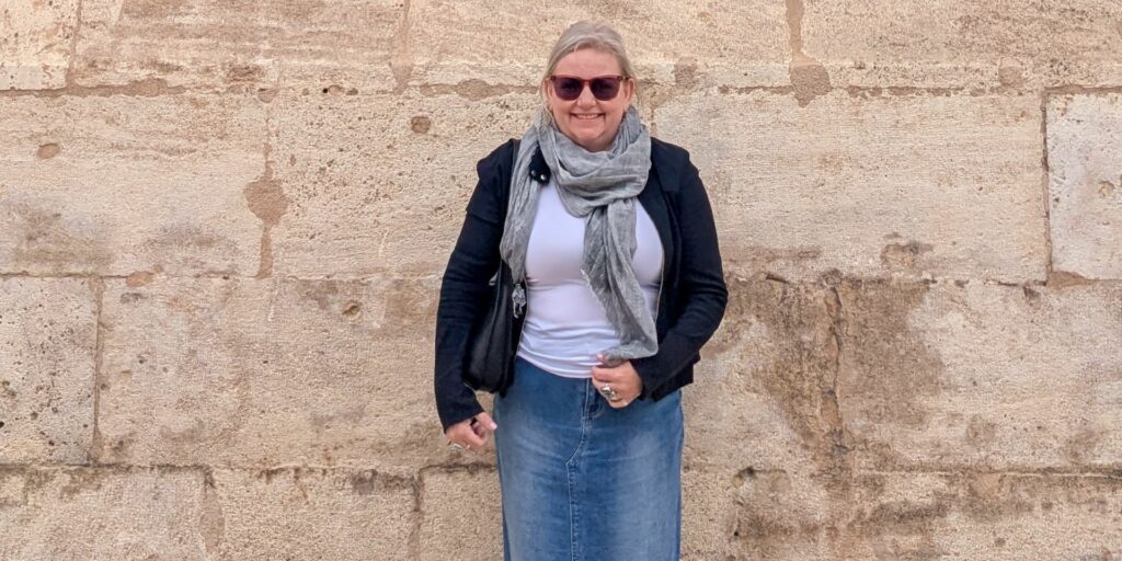Mell Millgate, of Mell Speaks, stands smiling in front of a textured sandstone wall, wearing sunglasses, a grey scarf, denim skirt, and a black jacket.