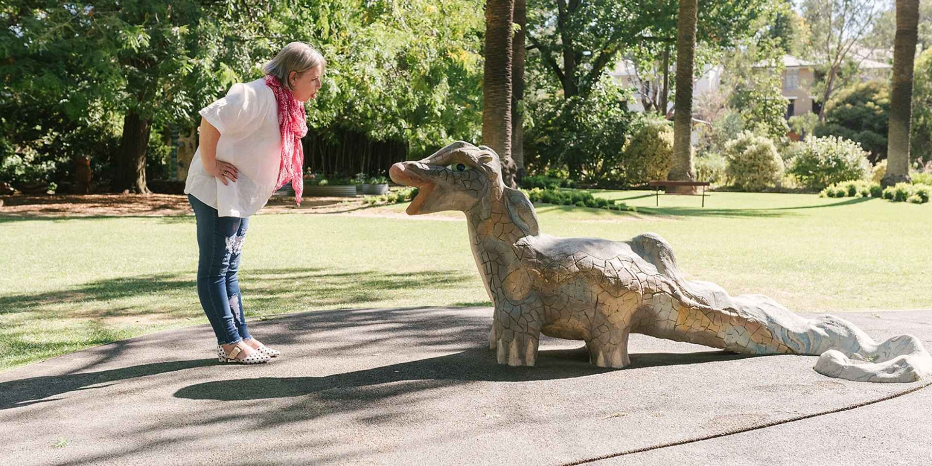 Woman in jeans and a white top leans forward playfully to face a mosaic-style dragon sculpture in a sunny park surrounded by green trees and grass.