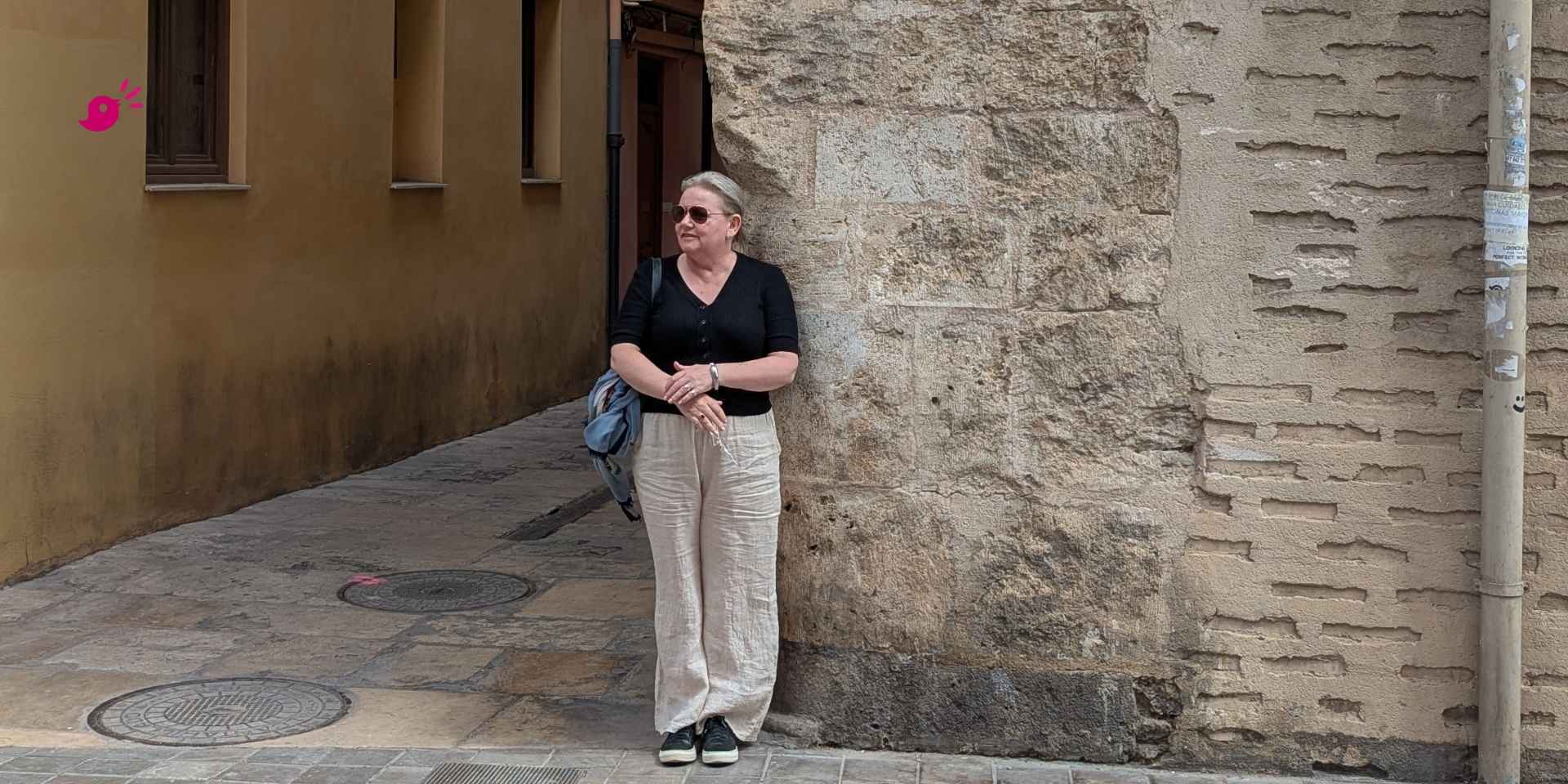 A woman stands in a narrow European street, leaning casually against a stone wall, wearing light linen trousers, a black top, sunglasses, and sneakers. She looks to the side with a calm, reflective expression.