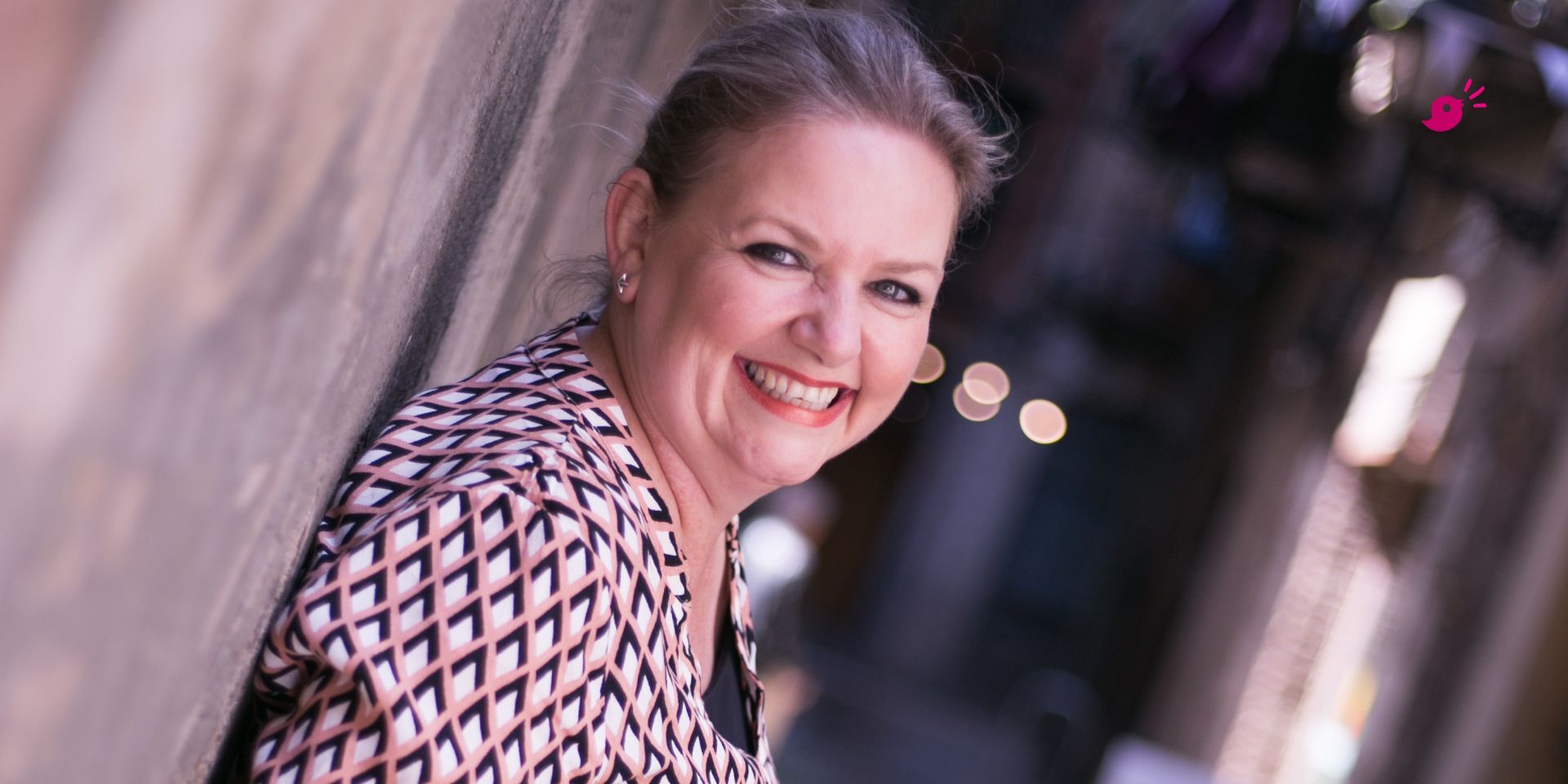 A smiling woman, Mell of Mell Speaks, with light hair pulled back is leaning against a wall in an outdoor laneway. She is wearing a patterned jacket and looking directly at the camera.