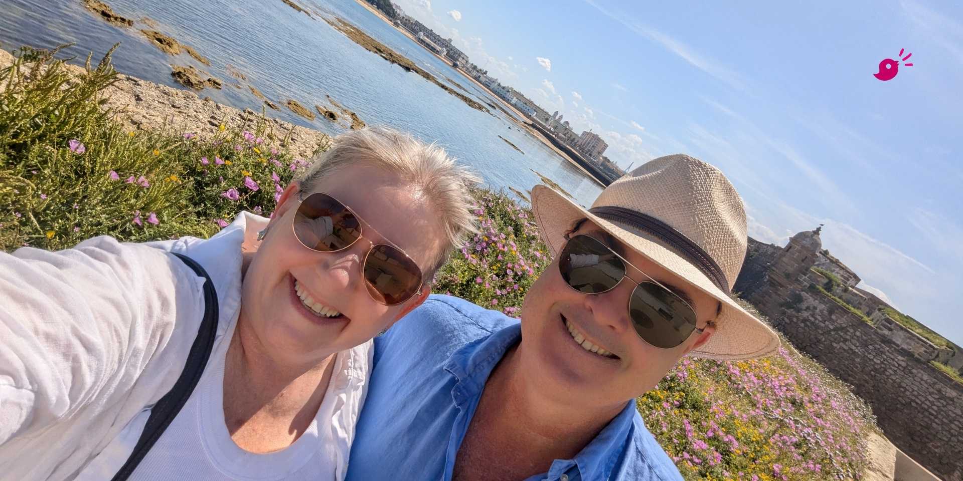 Mell and her partner Josh smiling at the camera beside the rocky coastline in Cádiz, Spain, with wildflowers, calm blue water and historic stone buildings behind them on a sunny day.