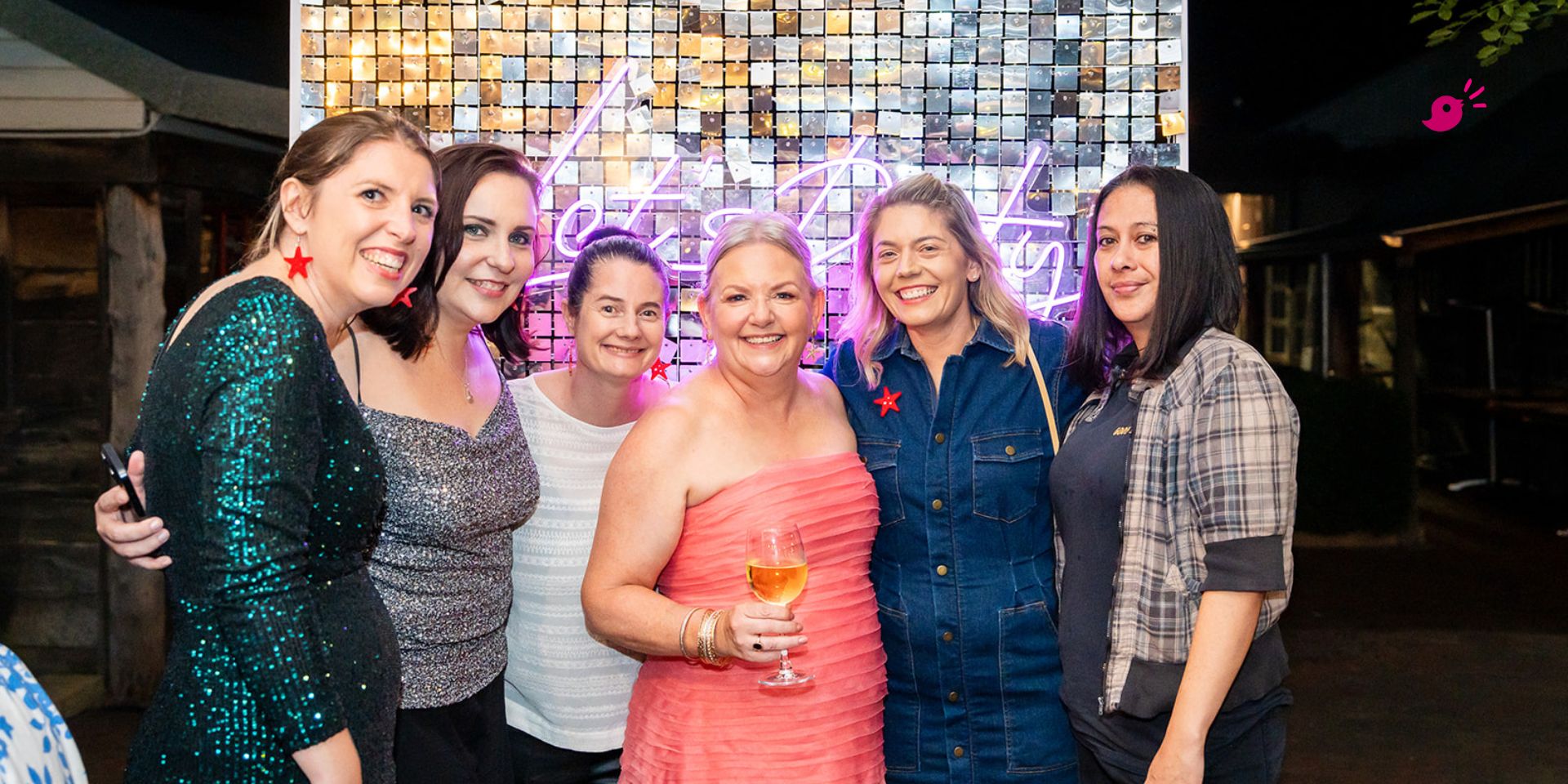 Group of six women at a business event smiling together in front of a lit backdrop, celebrating community and connection in business